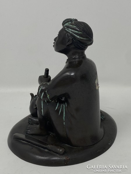 Marked bronze statue of an Indonesian boy sitting on the ground, smoking a pipe and drinking tea, c