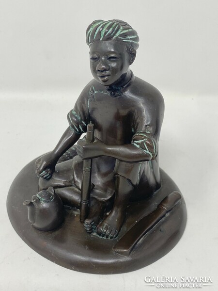 Marked bronze statue of an Indonesian boy sitting on the ground, smoking a pipe and drinking tea, c